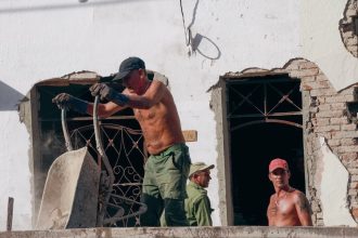 Três trabalhadores atuam em uma obra de reforma de um edifício, sob a luz do sol.