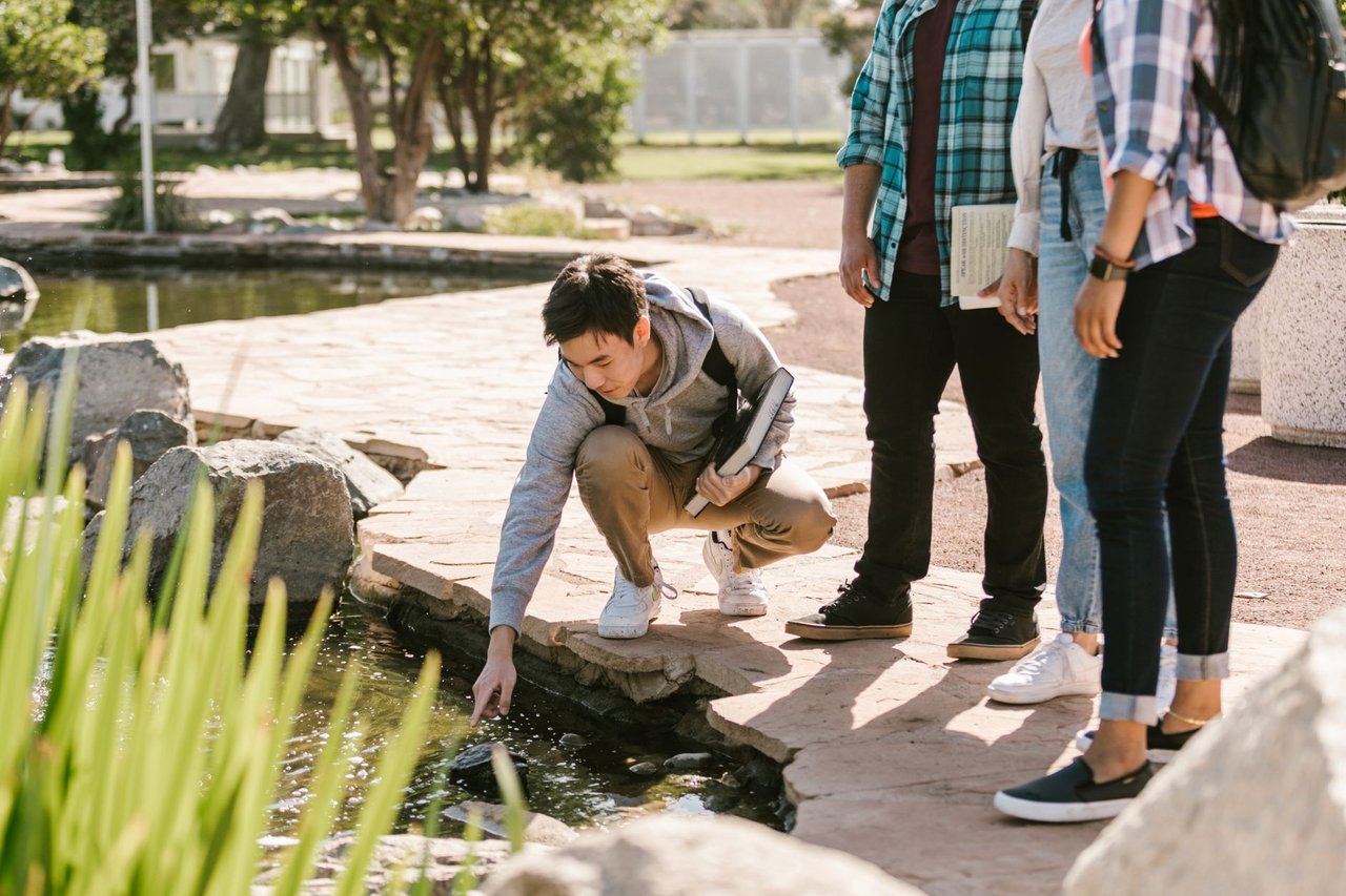 Grupo de estudantes participa de atividade de estudo junto a um lago, observando elementos da natureza.