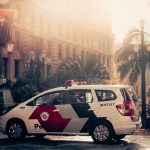 Viatura policial estacionada em uma via urbana, cercada por palmeiras e iluminada pela luz do dia.